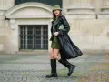 Kobieta w czarnym płaszczu i **oliwkowym** kapeluszu typu bucket hat, z zieloną torebką, na tle kamiennej fasady.