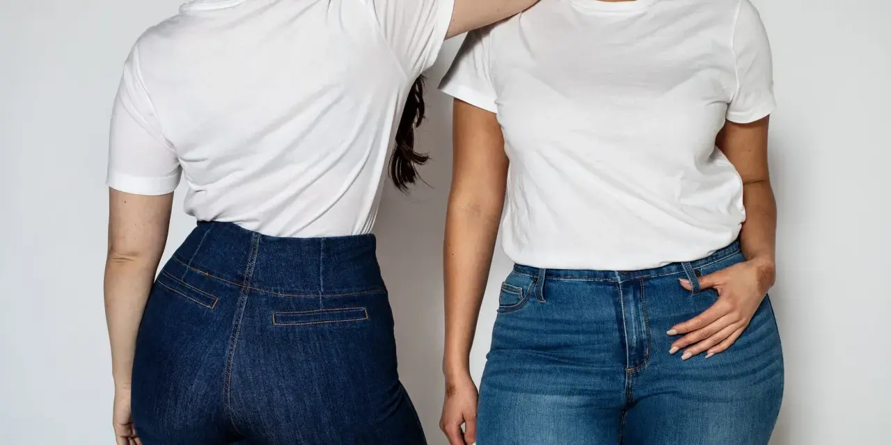 Dwie kobiety w białych T-shirtach i jeansach. Jedna stoi tyłem, druga przodem, opierając rękę na ramieniu koleżanki. Ich sylwetka v podkreślona przez dopasowane spodnie.