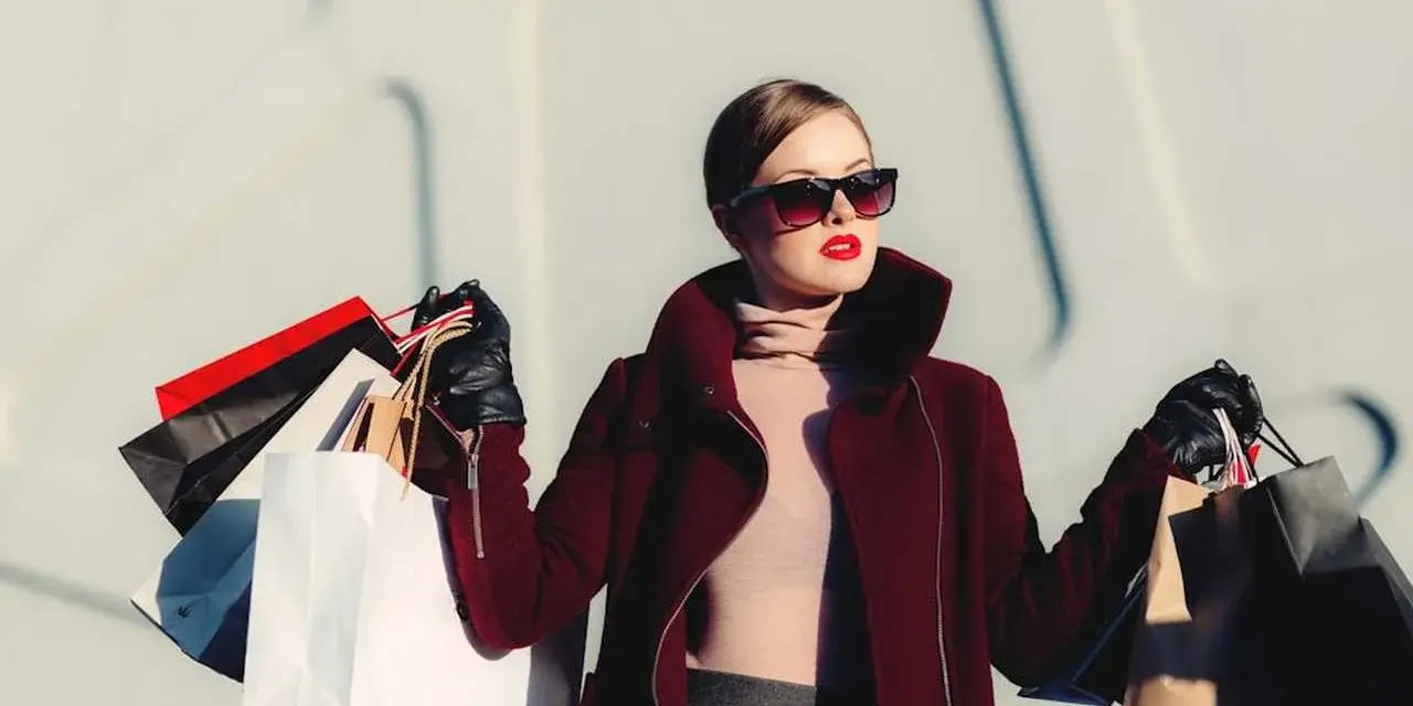 Elegancka kobieta z torbami zakupowymi, w okularach i czerwonym płaszczu. Jej styl to świetny przykład, jak można wyglądać modnie, co potwierdzają pozytywne dstreet opinie.