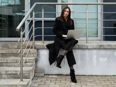 Elegancka kobieta w czarnym płaszczu i spodniach, z laptopem na kolanach, siedzi na schodach. Jej czarny płaszcz stylizacje podkreślają wyraziste buty i rajstopy w kropki.