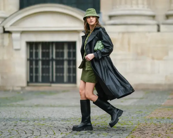 Kobieta w czarnym płaszczu i **oliwkowym** kapeluszu typu bucket hat, z zieloną torebką, na tle kamiennej fasady.