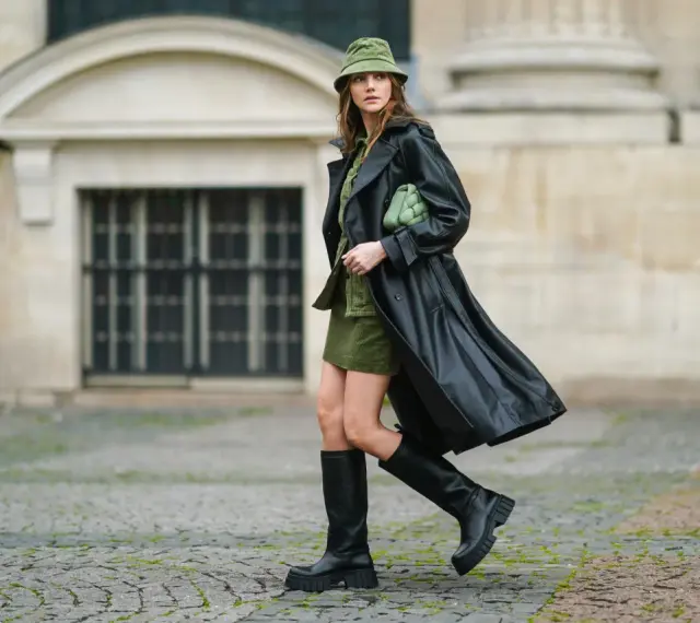 Kobieta w czarnym płaszczu i **oliwkowym** kapeluszu typu bucket hat, z zieloną torebką, na tle kamiennej fasady.