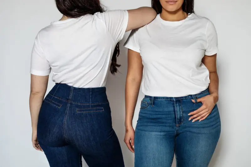 Dwie kobiety w białych T-shirtach i jeansach. Jedna stoi tyłem, druga przodem, opierając rękę na ramieniu koleżanki. Ich sylwetka v podkreślona przez dopasowane spodnie.