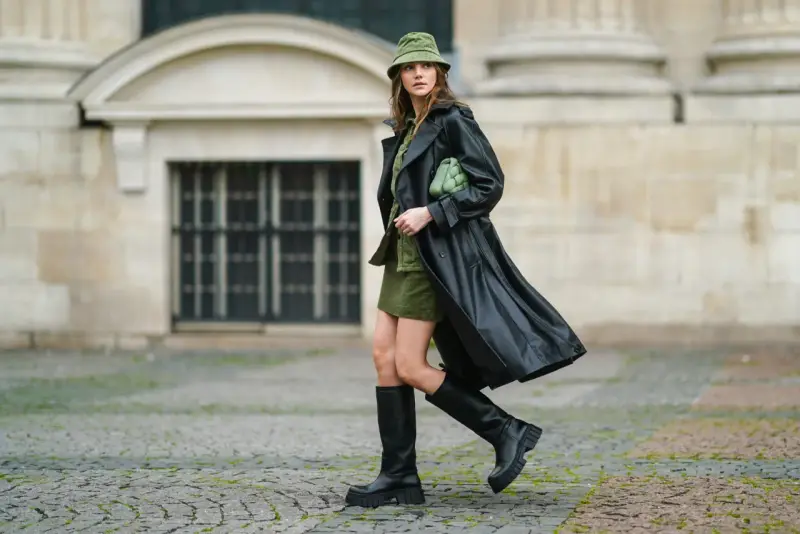Kobieta w czarnym płaszczu i **oliwkowym** kapeluszu typu bucket hat, z zieloną torebką, na tle kamiennej fasady.